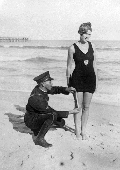 На пляже блюститель порядка измеряет даме длину купальника.
