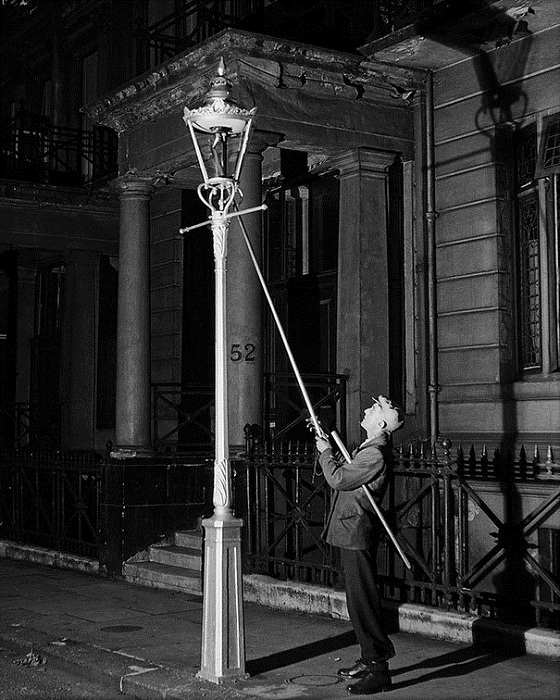 Фонарщик при помощи длинной палки зажигает уличный фонарь.