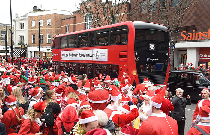Автобус едет сквозь огромную толпу людей, одетых в костюмы Санта Клауса. Несколько человек одеты в зеленые костюмы эльфов.