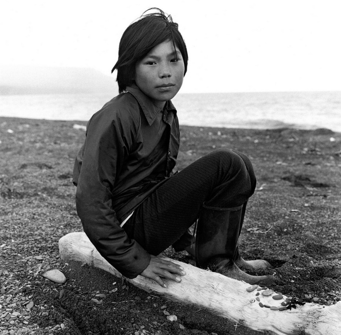 Ребенок в резиновых сапогах выкладывает из гальки узоры на бревне. Тунунак, Аляска, июль 1977 год.