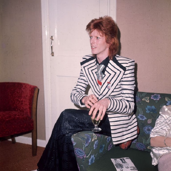 Дэвид Боуи в полосатом пиджаке с широкими лацканами и брюках-клеш с узором в виде блестящих квадратов.