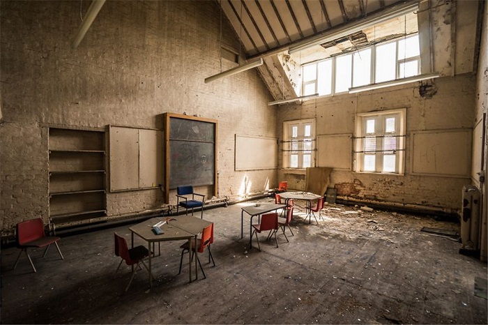 Покрытая слоем пыли грифельная доска соседствует со стоящими рядом столами и стульями в разрушающемся здании в Великобритании.