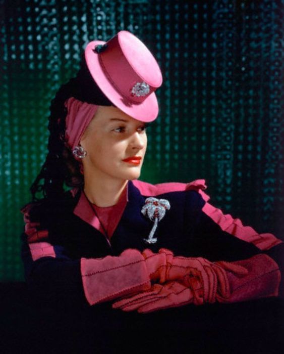 Модель в маленькой ярко-розовой шляпке с брошкой, сочетающейся с перчатками и вставками на костюме.