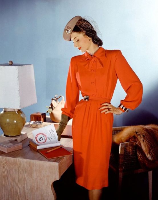 Модель в ярко-оранжевом платье с поясом и воротником-бантом, берет с сеточкой вместе с перчатками того же цвета дополняют образ.