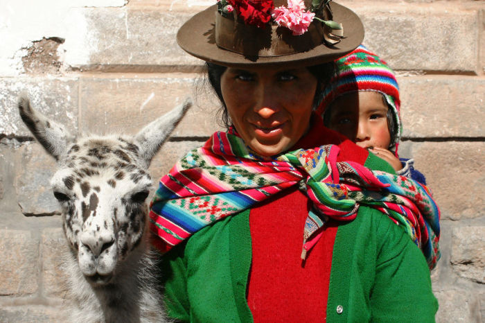 Мать в яркой одежде позирует вместе с сыном и альпакой на улице города, расположенного в Андах.