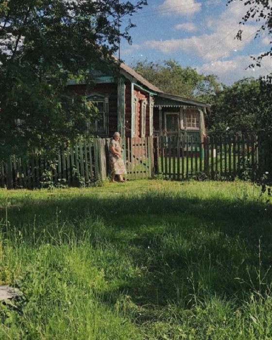 Пожилая жительница русской деревушки греется на солнышке возле своего двора.