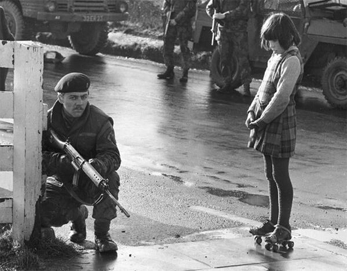 Девочка на роликовых коньках без страха смотрит на вооруженного британского солдата.