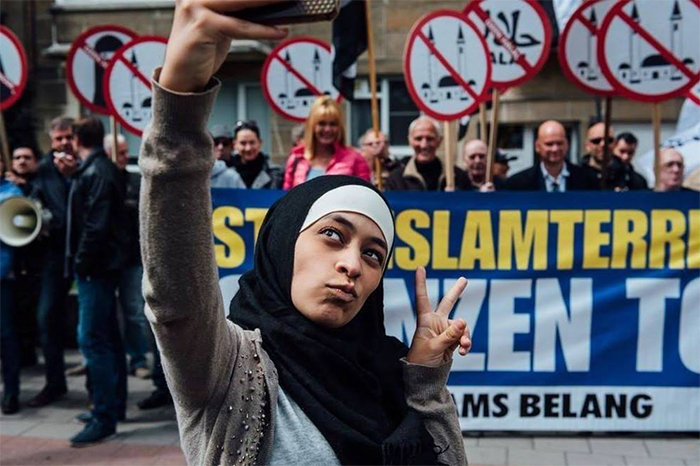 Закия Белхири делает селфи на фоне антимусульманской демонстрации, требующей запретить хиджабы и мечети.