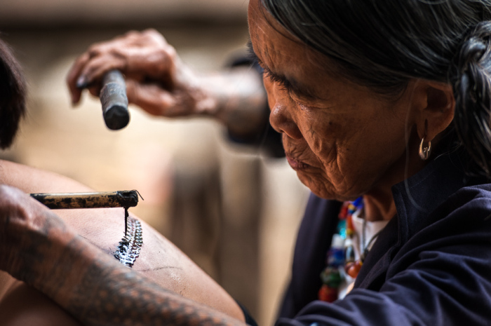 Фанг-од делает татуировку местному туристу из Манилы. Она использует шип дерева помело, прикрепленный к куску бамбука. В качестве чернил выступает сажа, которую она собирает на дне своей кастрюли.